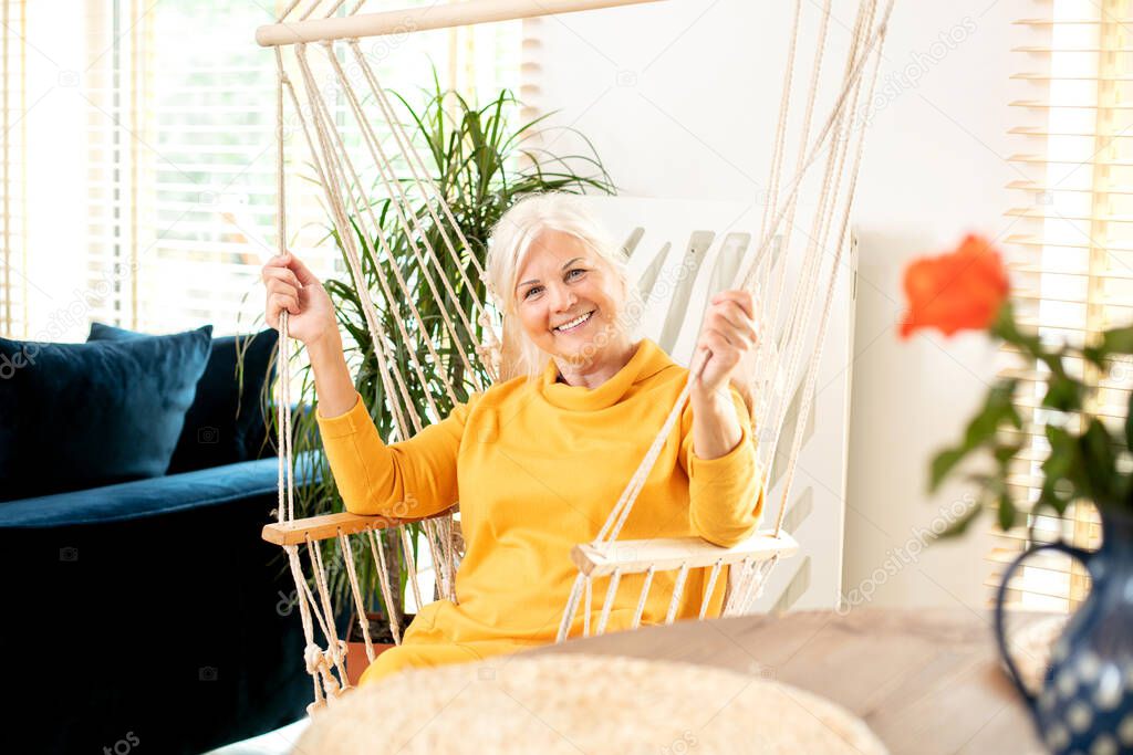 Portrait of beautiful senior woman sitting on a boho swing at home and smiling. Mature lady relaxing in living room. Happy retirement concept.