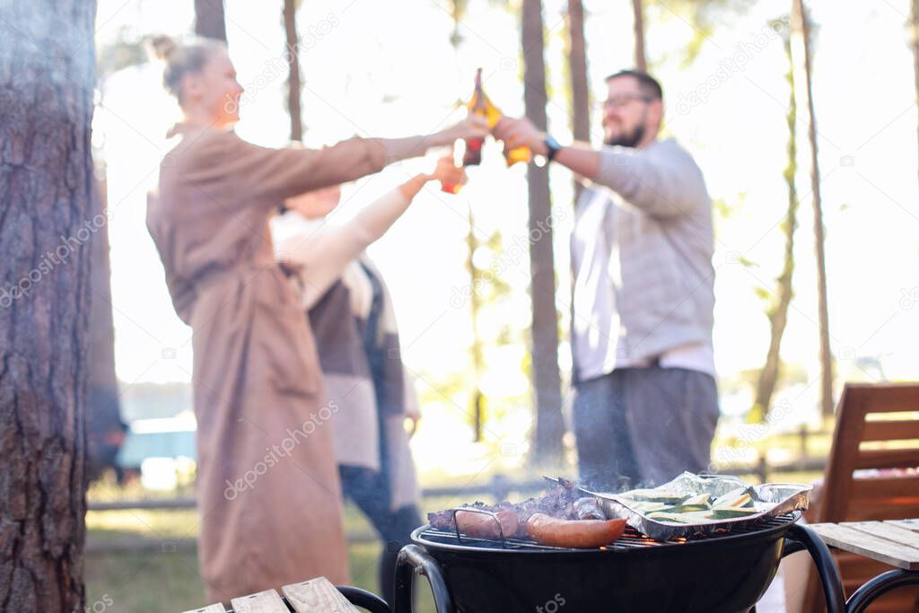 Group of friends having fun grilling meat and vegetables, enjoying bbq party. Happy guy and girls cooking and drinking beers at barbecue dinner outdoor, chatting.