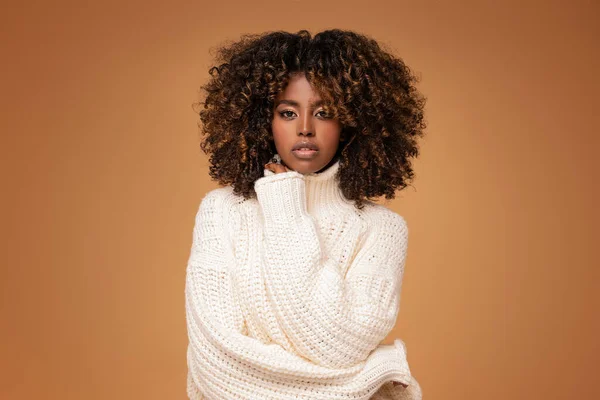 Happy beautiful african girl with afro hairstyle posing in cozy sweater on beige studio background.