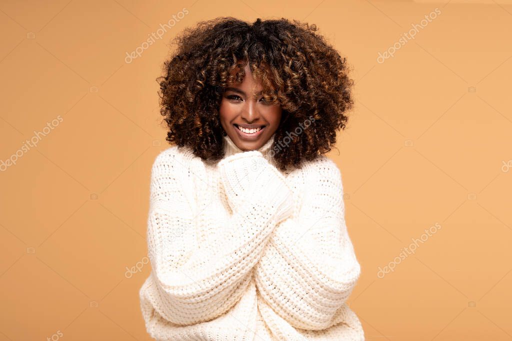Happy beautiful african girl with afro hairstyle posing in cozy sweater on beige studio background.