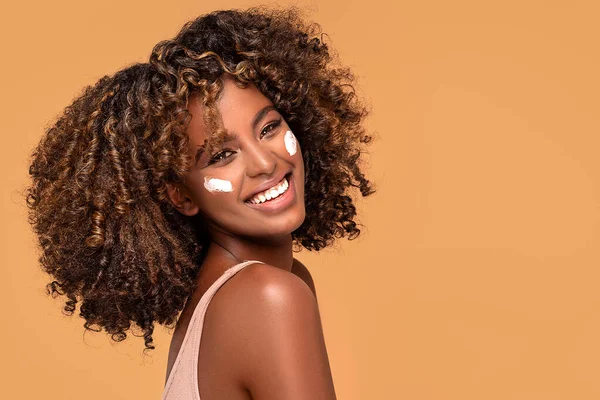 Yüzünde nemlendirici olan genç bir afro kadın. Güzel Afrikalı kız yanağına krem sürüyor. Mükemmel ve sağlıklı ciltli güzel kız..