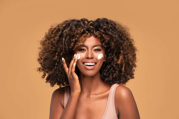 Young afro woman with moisturizer on face. Beautiful african girl applying a cream on cheek. Beauty girl with perfect and healthy skin.