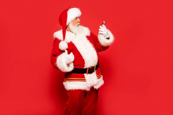 Real funny Santa Claus looking at mobile phone screen . posing over red background.