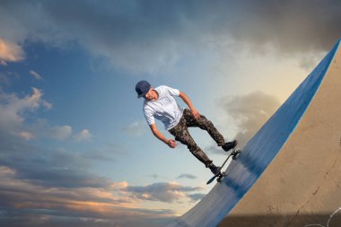 Genç kaykaycı gün batımında kaykayıyla bir rampadan iniyor.