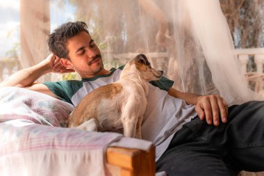 Gülümseyen genç bir adam sinekliğin altında köpeğinin yanındaki katlanır sandalyede oturur.