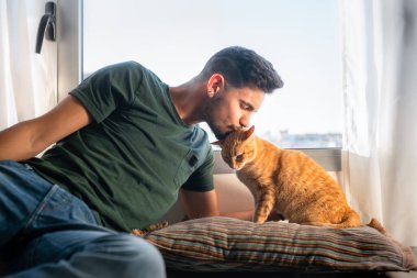 Genç adam pencerenin önünde tekir bir kedinin kafasını öpüyor.