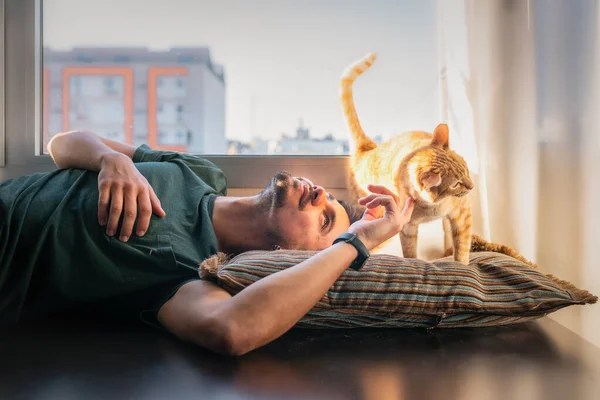 Молодой Человек Лежит Подушке Окном Гладит Шею Коричневой Кошки — стоковое фото
