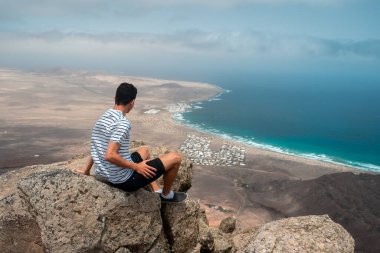 Uçurumun tepesinde oturan genç bir gezgin, arka planda Famara kasabasını düşünüyor. Teg giyin. Lanzarote. Kanarya Adaları