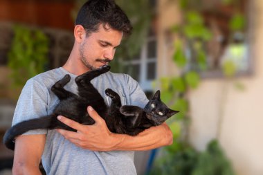Genç adam kollarında yeşil gözlü kara bir kedi tutuyor ve ona bakıyor. Kedi kameraya bakar.