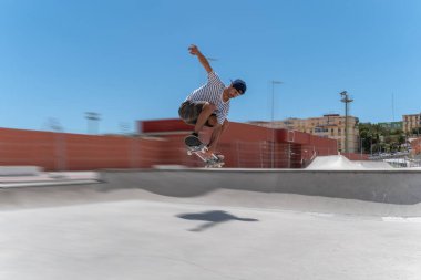 Genç adam güneşli bir günde kaykay parkında sörf tahtasıyla atlıyor.