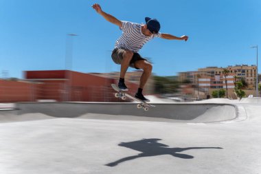 Genç adam güneşli bir günde kaykay parkında sörf tahtasıyla atlıyor.