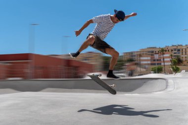 Genç adam güneşli bir günde kaykay parkında sörf tahtasıyla atlıyor.