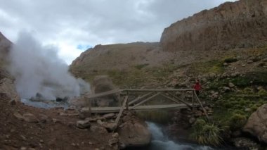 Yürüyüş ve Los Tachos Covunco Nehri üzerinde küçük ahşap pedestrean köprüden geçen Kırmızı montlu bir kadın. Geisers sıcak su ve arka plan üzerinde vaper. Neuquen, Patagonya Arjantin.