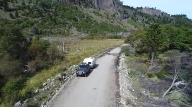 Sonbahar güneşli bir günde bir kamyonetle ıslak dirt road ve yeşil ağaçlar ile bozkır hava dron sahnesi. Araucarias bir taş duvar kamera hareketli ileriye atmak,
