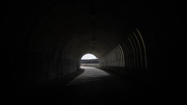 Hand held camera walking through and getting out a brutalist concrete tunnel. Lights and shadows rithms made by windows, openings, within the tunnel. Background of mountains, rocky walls and blue sky — Stock Video
