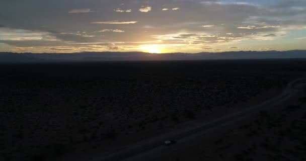 Escena aérea de drones de la hora dorada del atardecer con el sol en el horizonte sobre las montañas en el fondo con nubes. Cámara moviéndose hacia arriba. Ruta con furgoneta y casa remolque. La Rioja, Argentina . — Vídeos de Stock