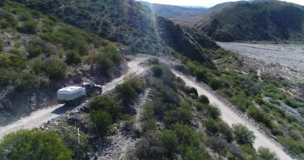 Luchtfoto drone scène van busje met aanhanger, reizen op onverharde weg aflopende hillslope van Famatina bergen. Camera volgt de 4 x 4 van achteruit. Vallei van de gele rivier en heuvels op de achtergrond. — Stockvideo