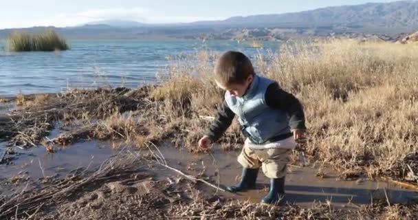 Ein Kind mit Regenstiefeln spielt im Schlamm am Ufer eines Sees und bewegt das schmutzige Wasser mit einem Baumbrunch. Hintergrund von See und Bergen. Staudamm Cuesta del Viento, Provinz San Juan, Argentinien — Stockvideo