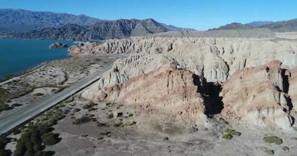 Scène aérienne de drone de lac bleu et de montagnes érodées avec des rivières et des ravins secs. Route le long de la côte de sable. En approche d'une colline rocheuse colorée. Barrage de Cuesta del Viento, province de San Juan — Video