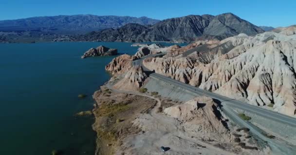 Letecká drone scéně létání nad pobřeží a silnici směrem na erodované bílé a oranžové pískovcové hory, modré jezero na boku. Černé a šedé hory v pozadí. Cuesta del viento dam — Stock video