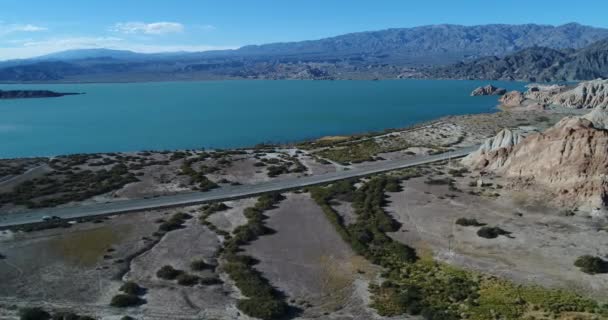 Drone aereo scena di lago blu e montagne erose con fiumi secchi e calanchi. Strada lungo la costa sabbiosa con vegetazione adattata alle condizioni saline. Avvicinamento costa e lago . — Video Stock