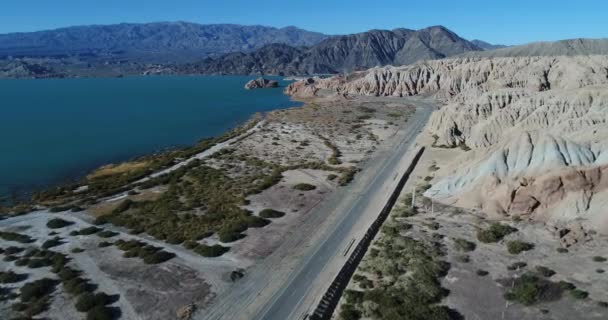 Cena de drones aéreos de lago azul e montanhas erodidas com rios secos e ravinas. Estrada ao longo da costa arenosa com vegetação adaptada às condições salinas. Cuesta del Viento Dam, província de San Juan — Vídeo de Stock