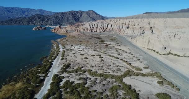 空中无人机现场飞越咸咸的海岸和道路 走向侵蚀的白色和橙色砂岩山脉 蓝色的湖在一边 背景是黑色和灰色的山脉 Cuesta Del Viento — 图库视频影像