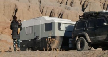 Kadın ve bebek karavana doğru gidiyor. Arka planda kuru turuncu dağlar var. Cuesta del Viento Barajı. San Juan Eyaleti, Arjantin