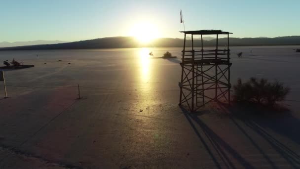 Escena aérea de drones de sillhuete de torre vigía de madera en plano desierto, puesta de sol. El sol de la hora dorada refleja sobre la superficie brillante de Barreal de Arauco, provincia de Rioja, Argentina. Infraestructura de los automóviles de vela — Vídeos de Stock