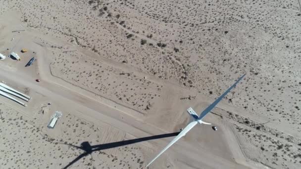 Scène de drone aérien vue de dessus senitale de l'ombre de moulage de turbine à treuil, machines travaillant sur de nouveaux moulins à vent avec des pales couchées sur le sol. Aimogasta, La Rioja, Argentine. Caméra panoramique vers le haut à l'horizon — Video