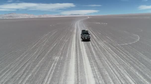 Escena aérea de aviones no tripulados siguiendo una furgoneta de expedición desde el frente conduciendo fuera de la carretera en el desierto gris. Montañas coloridas erosionadas en el fondo. Antofagasta de la Sierra, Catamarca, Argentina — Vídeo de stock