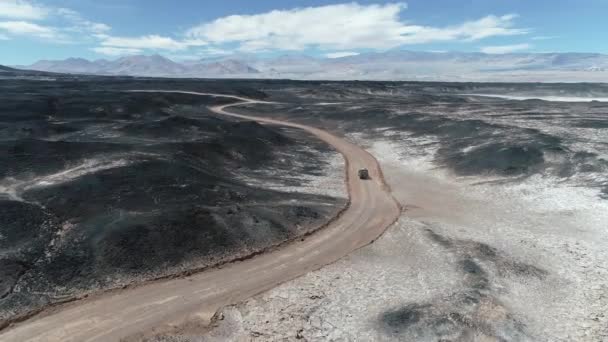 Escena aérea de camioneta de expedición 4x4 conduciendo fuera de la carretera entre lechos de lava, lapillis negros, región desértica oscura. Montañas coloridas al fondo. Paisaje árido místico. Catamarca, Argentina — Vídeo de stock