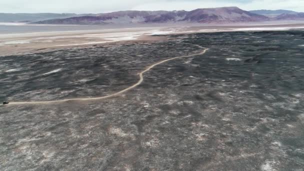 Escena aérea de camioneta de expedición 4x4 conduciendo fuera de la carretera entre lechos de lava, lapillis negros, región desértica oscura. Montañas rojas de colores al fondo. Paisaje árido místico. Catamarca, Argentina — Vídeos de Stock