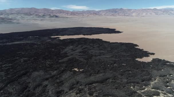 Volando sobre el lecho negro de lava. Montañas, paisaje desértico al fondo. Texturas volcánicas naturales, patrones. De vista frontal a senital. Antofagasta de la Sierra, Catamarca, Argentina — Vídeo de stock