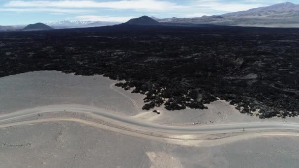 Fliegen sehr hoch an der Grenze zwischen schwarzem Lavabett und Wüste mit Straße. Wohnwagen, Auto mit Anhänger, kam auf dem Standstreifen zum Stehen. schwarze Vulkane im Hintergrund. antofagasta de la sierra, katamarca, argentina — Stockvideo