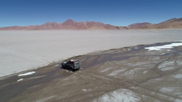 Escena aérea de drones de 4x4 conduciendo fuera de la carretera cruzando un río y viajando sobre un amplio desierto. Montañas coloridas en el fondo del cráter del volcán Galán. Antofagasta de la Sierra, Catamarca, Argentina — Vídeo de stock