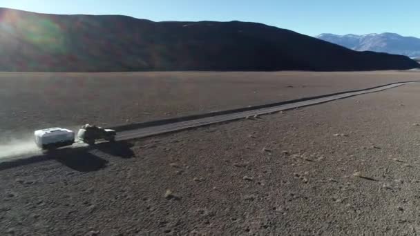 Escena aérea de drones de 4x4 con caravana que viaja fuera de la carretera en pista de grava en montañas desérticas. Siguiendo el coche desde el costado. Bengalas y luz de volumen. Andes al fondo. Antofalla, Catamarca — Vídeo de stock