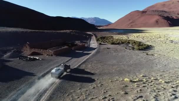 Drohnen-Szene eines Lieferwagens mit Wohnwagen, der über einen Schotterweg zwischen verlassenem Lehmhaus und verlassenem Tal fährt. salar und erodierten Bergen im Hintergrund. katamarca, argentinien — Stockvideo