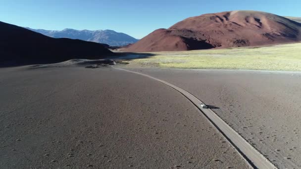 Escena aérea de drones de furgoneta con caravana que viaja fuera de la carretera en la pista de grava en el valle de la montaña desértica. Vista general del paisaje acercándose coche por detrás. Andes al fondo. Antofalla — Vídeos de Stock