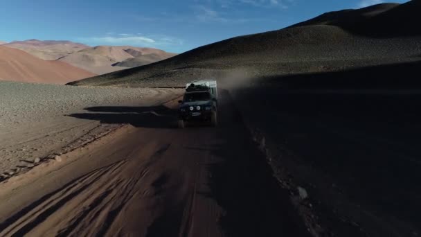 Aérea siguiendo desde el frente una camioneta de expedición con caravana que viaja fuera de la carretera en la pista de grava en las montañas de colores desérticos al atardecer. Antofalla, Catamarca, Argentina — Vídeo de stock