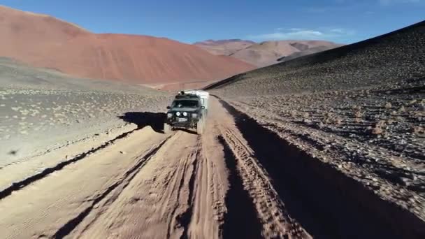 De cerca siguiendo desde el frente una camioneta de expedición con caravana que viaja fuera de la carretera en la pista de grava en las montañas desérticas al atardecer. Antofalla, Catamarca, Argentina — Vídeo de stock