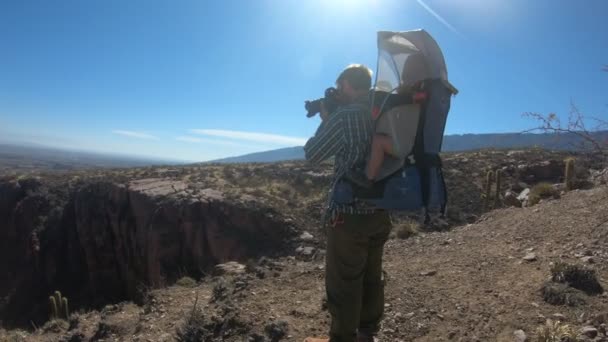 Ünlü fotoğrafçı, oğluyla birlikte kayalık kanyonun geniş bir manzarasında sırt çantasıyla yürüyüş yaparken, yan yana kameralar çekerken. Quebrada de Hualco, San Blas, Rioja Eyaleti, Arjantin. — Stok video