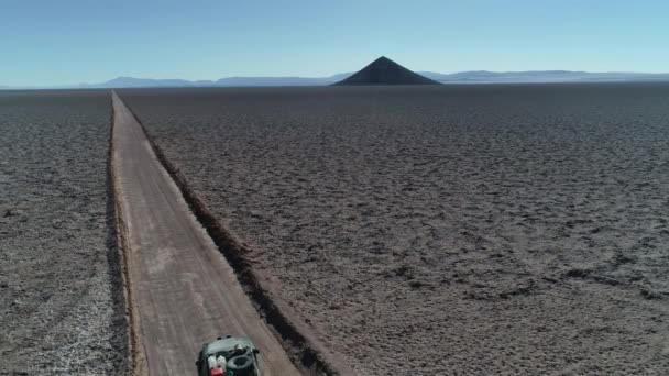 Αεροκατευθυνόμενα σκηνή 4x4 με τροχόσπιτο που ταξιδεύει εκτός δρόμου σε πλατιά salar με φυσική πυραμίδα, Arita Cone. Ορεινές αλυσίδες στο παρασκήνιο. Αριάρο Σαλάρ, Σάλτα, Αργεντινή. — Αρχείο Βίντεο