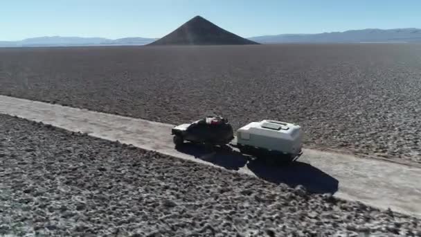 Cena de drone aéreo seguindo do lado muito perto 4x4 com caravana viajando fora da estrada em salar largo com pirâmide natural no fundo. Correntes de montanha no fundo. Arita cone, Arizaro salar — Vídeo de Stock