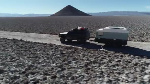 Following from the side a 4x4 with caravan traveling off road on wide salar with natural pyramid at background. Flying over car, general view of landscape. Arita cone, Arizaro salar — Stock Video