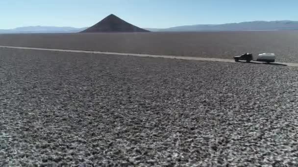 Antena następujące z boku 4x4 z przyczepą kempingową podróży Off Road na szerokim Salar z naturalnej piramidy w tle. Łańcuchy górskie w tle. Arita stożek, Arizaro Salar, Salta — Wideo stockowe