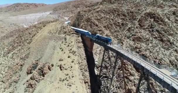 Aerial flying around train while it drives through old metal bridge at desertic mountainous landscape. General view of iron bridge. Train of the couds, tren de las nubes, San Antonio de los Cobres — Stock Video