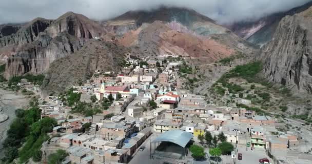 Voar aéreo sobre Iruya, cidade escondida, em vale estreito íngreme cercado de nuvens, bosques e colinas coloridas secas no fundo. Cidade turística em Salta, Argentina — Vídeo de Stock