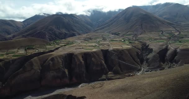 Ogólna antena gospodarstw rolnych przywieszanie od górskich zboczach ograniczających w kierunku głębokiego urwiska. Chmury i wysoki łańcuch górski w tle. Pueblo Viejo, Salta, Argentyna — Wideo stockowe