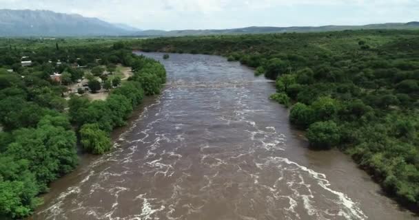 Scena z dronami latającymi do tyłu nad brązową rzeką przepływającą przez most. Powódź w Mina clavero. Rosnąca rzeka nad ulicami miasta. Kordoba, Argentyna — Wideo stockowe